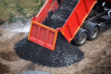 Red dump truck unloading crushed stone material onto construction ground, vehicle discharging bulk aggregate onto prepared site, heavy machinery performing roadwork material delivery © HENADZY