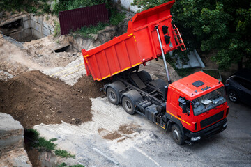 Red dump truck actively unloads pile of brown soil at urban construction site. Large heavy vehicle with raised bed deposits dirt near excavation pit and concrete structures © HENADZY