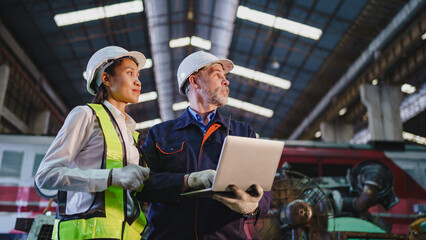 Fototapeta premium Industrial worker and engineer wearing helmet, safety vest, and glove while reviewing laptop inside manufacturing facility