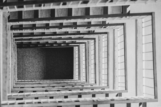 Black and white top-down view of a multistory square stairwell with repeating railings and steps, creating geometric perspective, deep symmetry and an abstract architectural pattern.