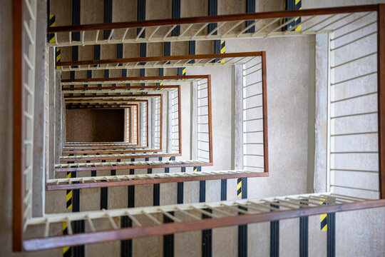 Top-down view into a multistory square stairwell with repeating stairs and railings, creating strong geometric perspective, depth, symmetry and an abstract architectural pattern.