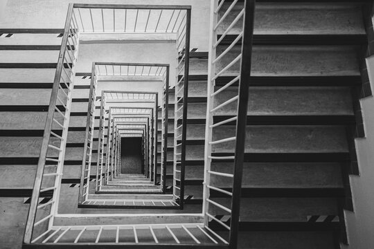 Black and white top-down view of a multistory square stairwell with repeating stairs and railings, creating strong geometric perspective, symmetry, depth and an abstract architectural pattern.