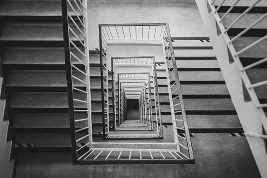 Black and white top-down view of a multistory square stairwell with repeating stairs and railings, creating strong geometric perspective, symmetry, depth and an abstract architectural pattern.