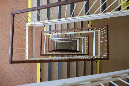 Top-down view into a multistory square stairwell with repeating stairs and railings, creating a striking geometric perspective, optical illusion, symmetry, depth and infinity tunnel effect.