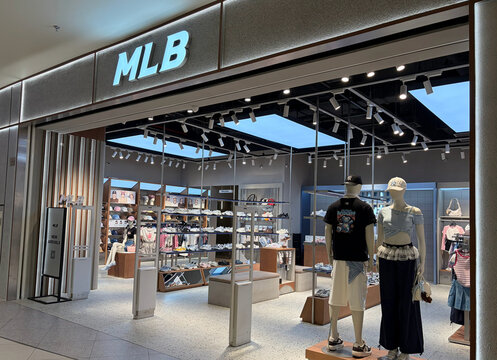 A modern retail store with the MLB logo prominently displayed. The store features stylish clothing and accessories. Mannequins showcase outfits, and shelves are neatly organized. The interior is well-