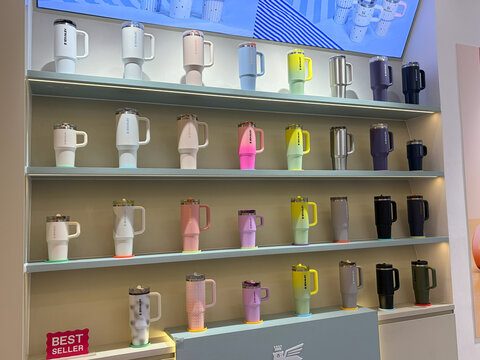 A row of colorful mugs displayed on shelves in a modern store environment. The mugs come in various shades and styles, featuring a range of designs. They are arranged neatly for visual appeal.