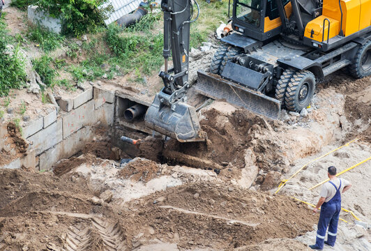 Excavator bucket digs earth trench for foundation work. Construction machinery moving soil during utility installation or building site preparation. Worker observes digging process 