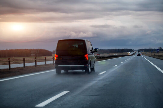 Rear view of black passenger van moving on multilane highway during evening hours, vehicle driving on wet asphalt road surface under cloudy sky, road transportation concept, evening sunset. 