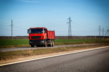Bright red dump truck travels on asphalt highway road through rural landscape side by side lush green field clear blue sky overhead power lines and utility poles © HENADZY