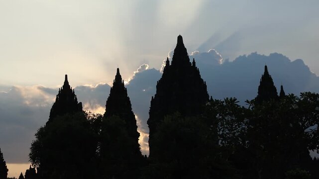 Silhouetted temple peaks reach toward a radiant sky. Sunlight spills through clouds, casting sacred rays. Ancient architecture stands still against a quiet horizon. Mystical glow suggests reverence