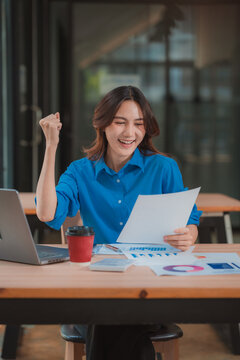 Excited Asian businesswoman raising arm celebrating business success while holding document at office desk