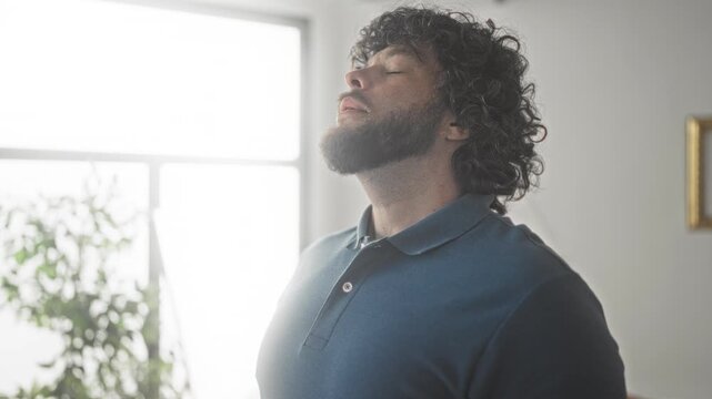 Man with beard and curly hair tilting head back and closing eyes beside sunlit window, wearing blue polo in studio space; quiet relief.