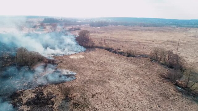 Dramatic aerial drone shot flying over a vast, burning dry grassland, large plumes of thick smoke billow into the sky as the wildfire spreads across the rural landscape