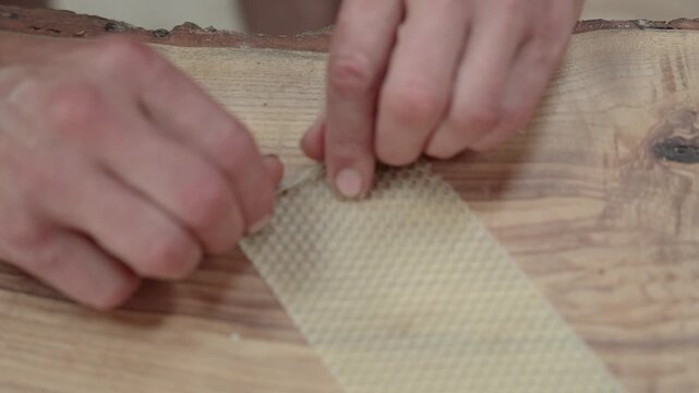 Close-up of hands positioning a wick along a textured beeswax sheet on a rustic wooden surface during handmade candle making