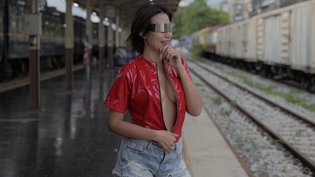 Asian woman wearing futuristic metallic eyewear, a red vinyl top, and denim shorts stands on a train station platform beside railway tracks