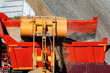 Aerial view of excavator bucket filling large red dump truck with sand material at construction site, heavy machinery performing earth moving work in bright daylight, copy space © HENADZY