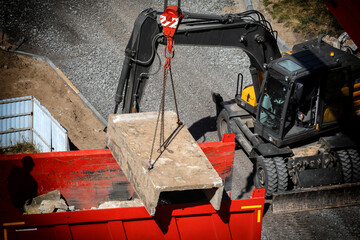 Heavy construction machinery operating on building site, excavator-loader lifting large concrete slab with crane attachment and chains, lowering into red dump truck bed, high angle view day outdoor © HENADZY