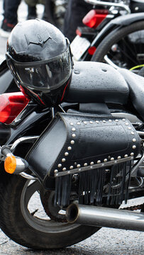 Black motorcycle parked on wet asphalt, close up detail of helmet and studded saddlebag with fringe after rain, chrome exhaust pipe and orange turn signal, vertical composition
