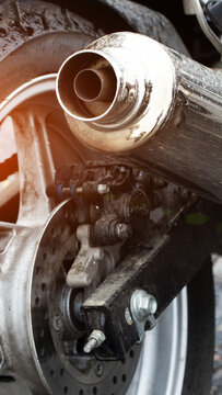 Close-up detail shot of motorcycle exhaust pipe and rear wheel assembly featuring disc brake, swingarm, and muddy tire tread, highlighting road dirt and water droplets on metal surface