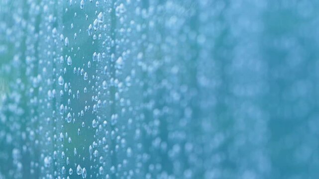 Extreme close up of water droplets on a window pane during a rainstorm. Authentic nature scene with soft blue tones for wellness and sustainability themes.