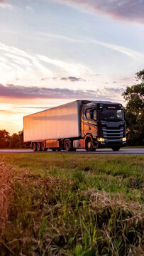 Black semi-truck with white cargo trailer driving on rural highway during colorful sunset, warm light reflecting on vehicle and sky, trucking industry transportation vertical photo