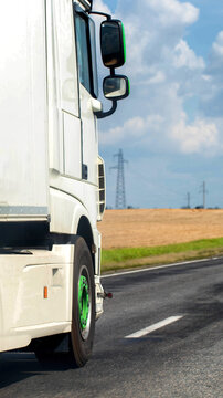 Side view of white semi-truck moving on asphalt road, green wheel hub, power lines and fields in background under blue sky with clouds, symbolizing cargo transportation logistics vertical photo