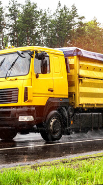 Yellow heavy duty truck with large cargo body driving on wet asphalt road during rain, water splashing from wheels, green roadside vegetation and blurred trees background, vertical photo