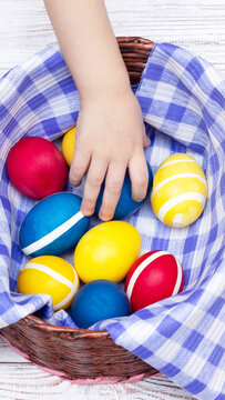 Child hand reaching for vibrant red blue and yellow Easter eggs with white stripes arranged in wicker basket lined with blue and white checkered cloth on white wooden surface vertical photo