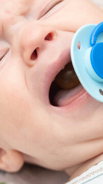 Delicate close-up portrait of serene sleeping infant face, pink cheeks, soft skin, blue pacifier providing comfort in open mouth, quiet moment. vertical photo