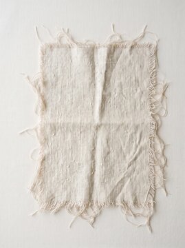Frayed Linen Fabric Swatch Flat Lay. A square piece of natural woven linen fabric with frayed edges lies flat on a plain light gray background.