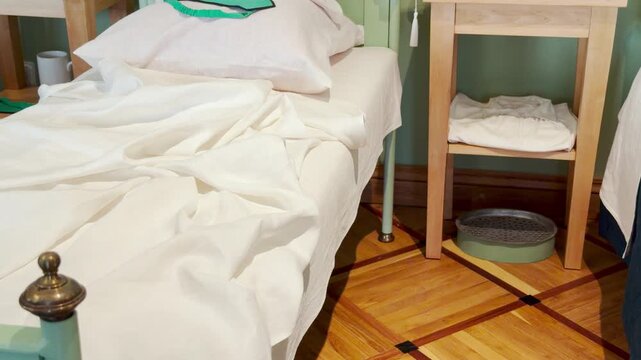 A bed with white sheets and slippers rests on a parquet floor in the Lyceum hospital ward in Pushkin, Saint Petersburg. The quiet scene brings a gentle feeling of nostalgic peace.