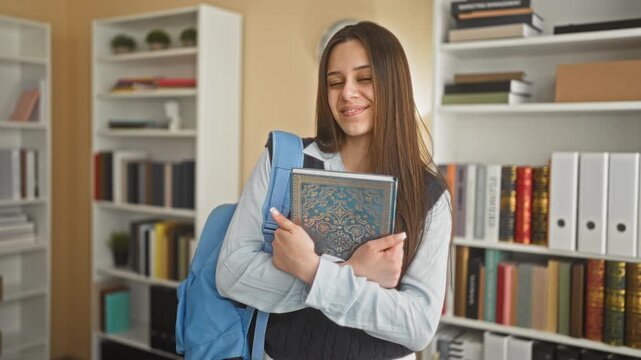 Woman student holding patterned book to chest, smiling while wearing backpack in library building among bookshelves and binders; confidence study.