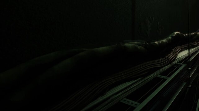 Cables run along a wall in a dimly lit underground area. The light creates shadows that add to the mystery of the scene. The setting feels isolated and hidden from view.