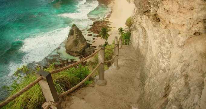 Stone stairs lead down steep cliffside toward sandy beach and bright ocean waves in Nusa Penida Bali