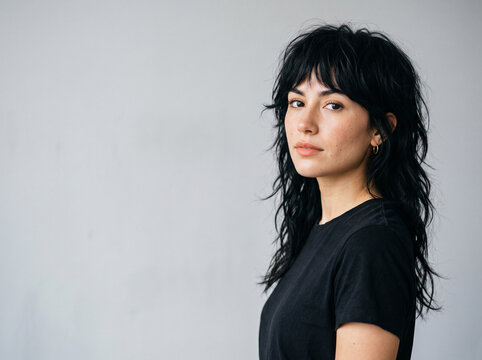 Retrato de mujer joven latina con cabello negro rizado estilo mullet posando sobre fondo gris. Peinados y cortes de pelo modernos y tendencias actuales en peluquer&iacute;as. Modelo con pelo negro flequillo