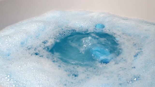 hand dropping a blue bath bomb into soapy water creating bubbles, foam fizzing, spa relaxation and wellness routine