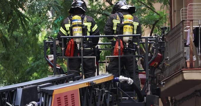 A fully equipped firefighting crew ascending an aerial ladder toward a burning building. The footage captures the firefighters in full gear, the extending mechanism of the ladder