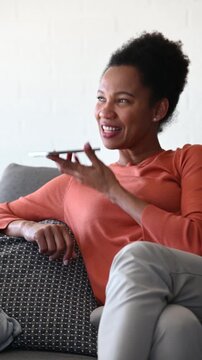 Black woman using smartphone to send messages and record a voice message while relaxing on sofa at home. Casual mobile communication and connected lifestyle