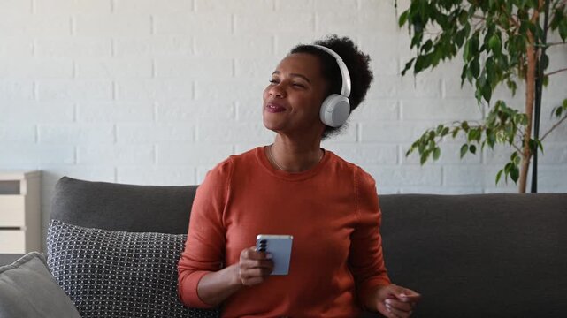 Happy Black woman wearing headphones, listening to music and dancing with smartphone while relaxing at home. Joyful lifestyle moment