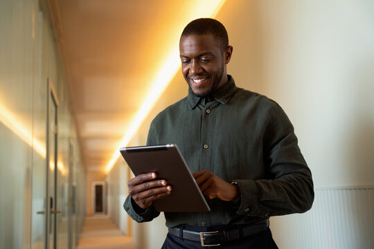 African 30s business man using digital tablet in office corridor, smiling black professional browsing online reports. Focused manager checks company data on touchscreen device. Copy space, employees.