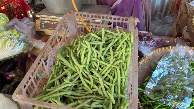 A video of a vegetable vendor selecting fresh green beans from a market stall. Their hands move gently, touching the vegetables. A plastic crate holds a large quantity of long, bright green beans. 4K 