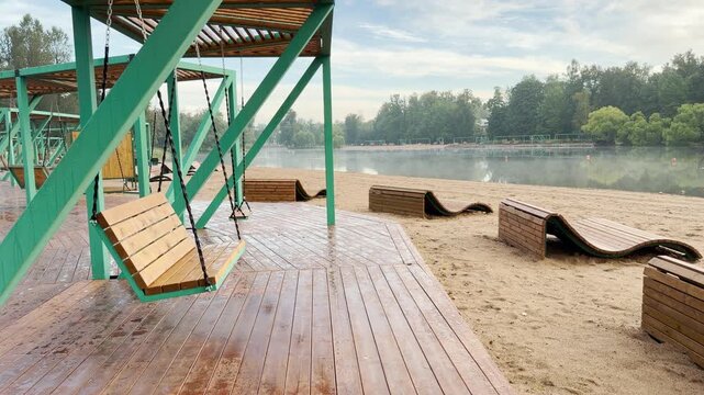 Wooden swings and loungers invite relaxation on a serene sandy beach by the calm Kolonistsky Pond lake in Pushkin, Saint Petersburg. This peaceful public space evokes a tranquil, relaxing feeling.