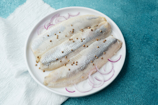 Marinated herring fillets with coriander seeds, onion and herbs on white plate with napkin over blue background