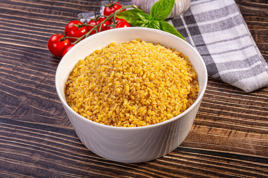 Bowl of golden bulgur grains on dark wood table with cherry tomatoes, basil, and garlic in background, rustic kitchen setting
