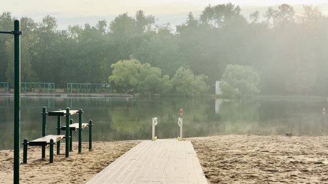 A wooden boardwalk leads toward a misty lake on a sandy beach in Pushkin, Saint Petersburg. This peaceful morning landscape evokes a calm, tranquil feeling.