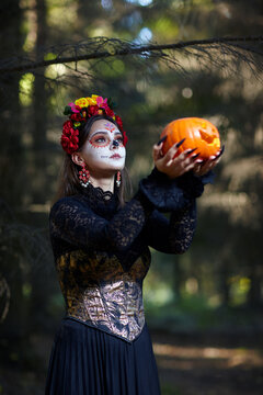 Woman with Santa Muerte face make-up in the forest holds a Halloween pumpkin in outstretched arms.