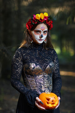 Halloween beauty portrait of woman in forest with Halloween pumpkin in hand. Santa Muerte makeup.