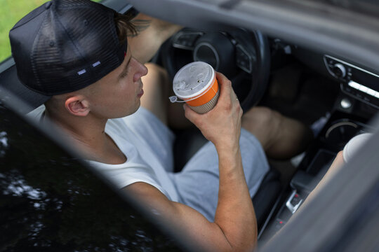 High-angle view of a young man in a baseball cap driving a car and taking a sip from a paper takeaway coffee cup. A modern, casual morning commute or road trip