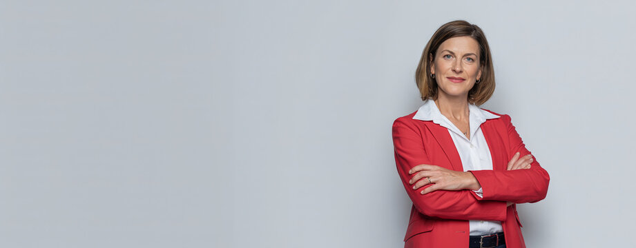 Confident businesswoman in red blazer with arms crossed against gray background.