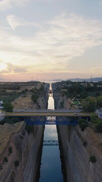 Vertical drone above Corinth Canal, Greece at sunset. Bridges span narrow waterway, cars crossing. Few clouds, clear sky. Golden light